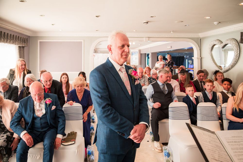Wedding guests watch and smile during an emotional moment at an indoor wedding ceremony with white chairs and decor.