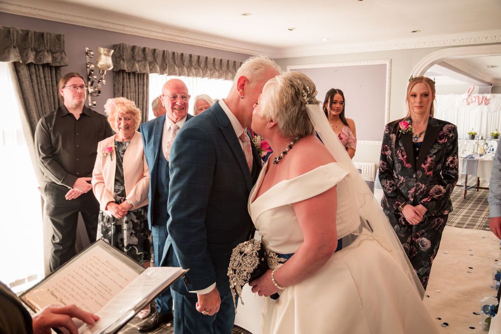 A couple shares their first kiss during an intimate wedding ceremony while guests watch in a bright indoor venue.