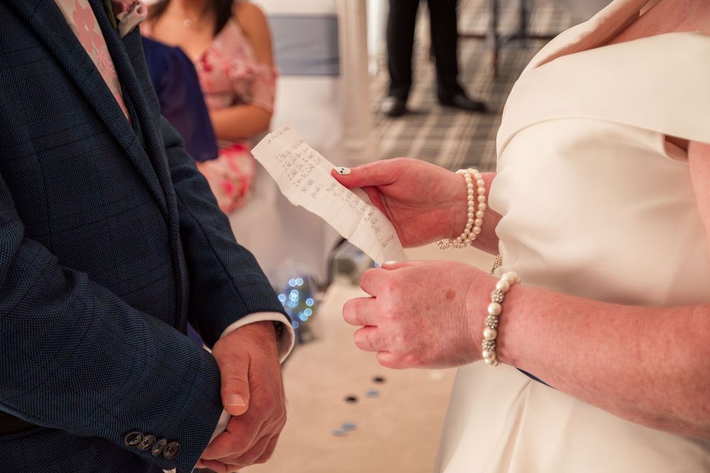 Close-up of hands holding wedding vows during ceremony with bridal jewelry and formal attire visible.