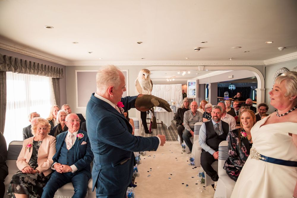At a wedding ceremony, a celebrant in a navy suit leads the proceedings as guests watch from their seats.