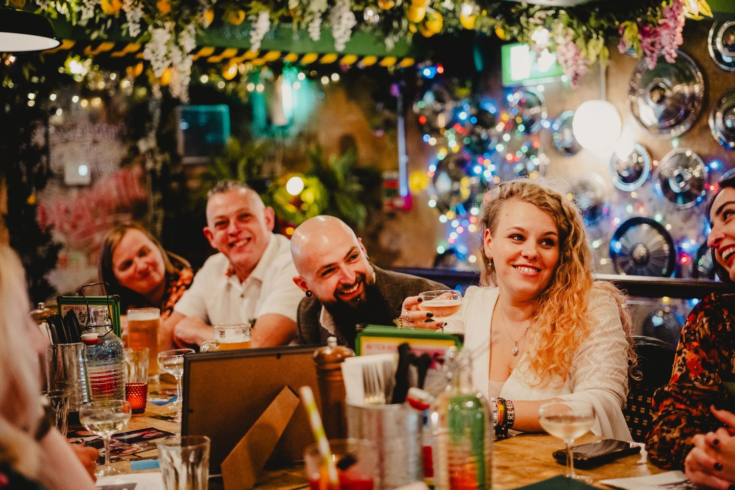 Friends enjoy a festive dinner party with drinks under decorative string lights and Christmas decorations.