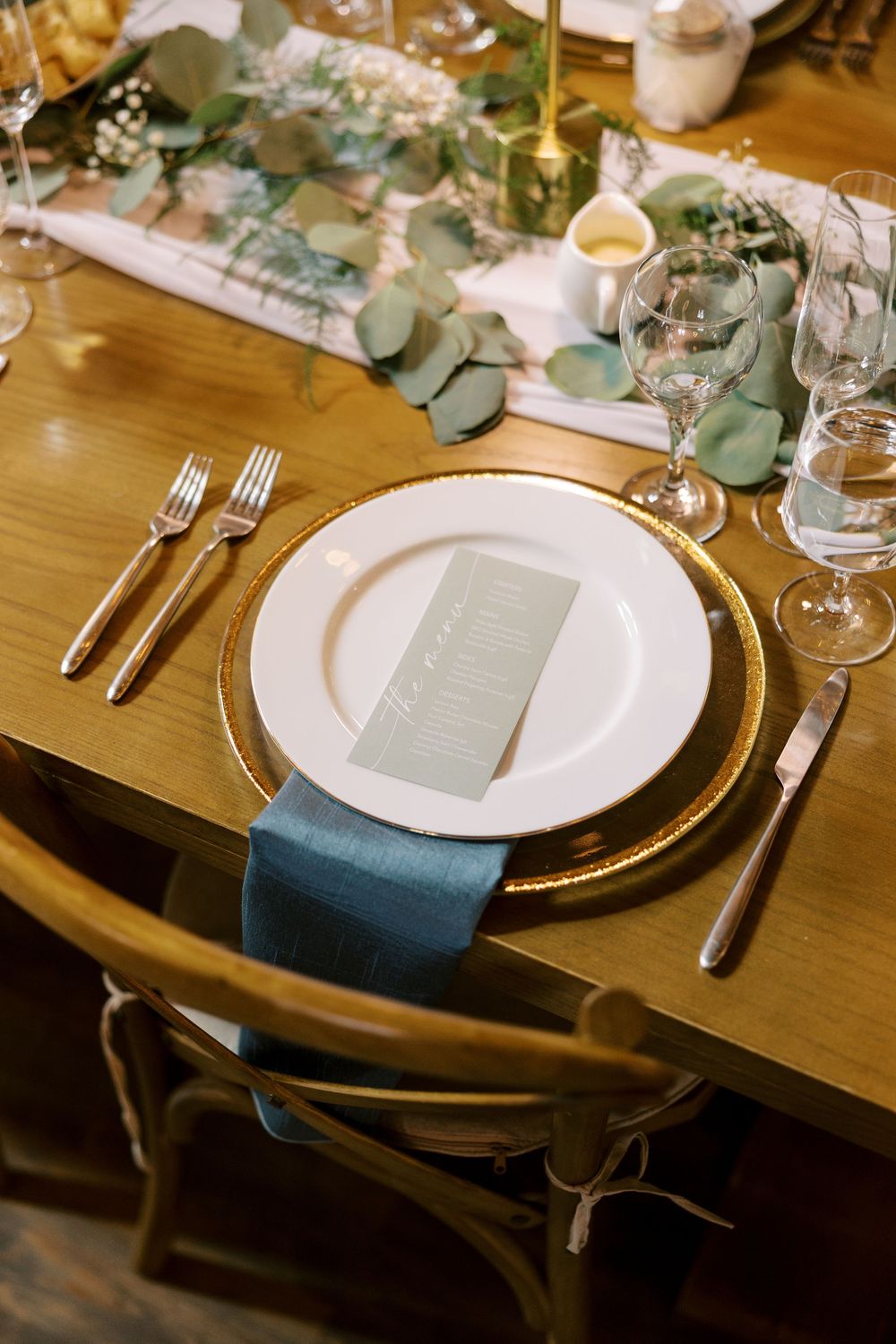Elegant table setting with gold charger plate, teal napkin, eucalyptus garland, and menu card on wooden farm table at Windham Manor.