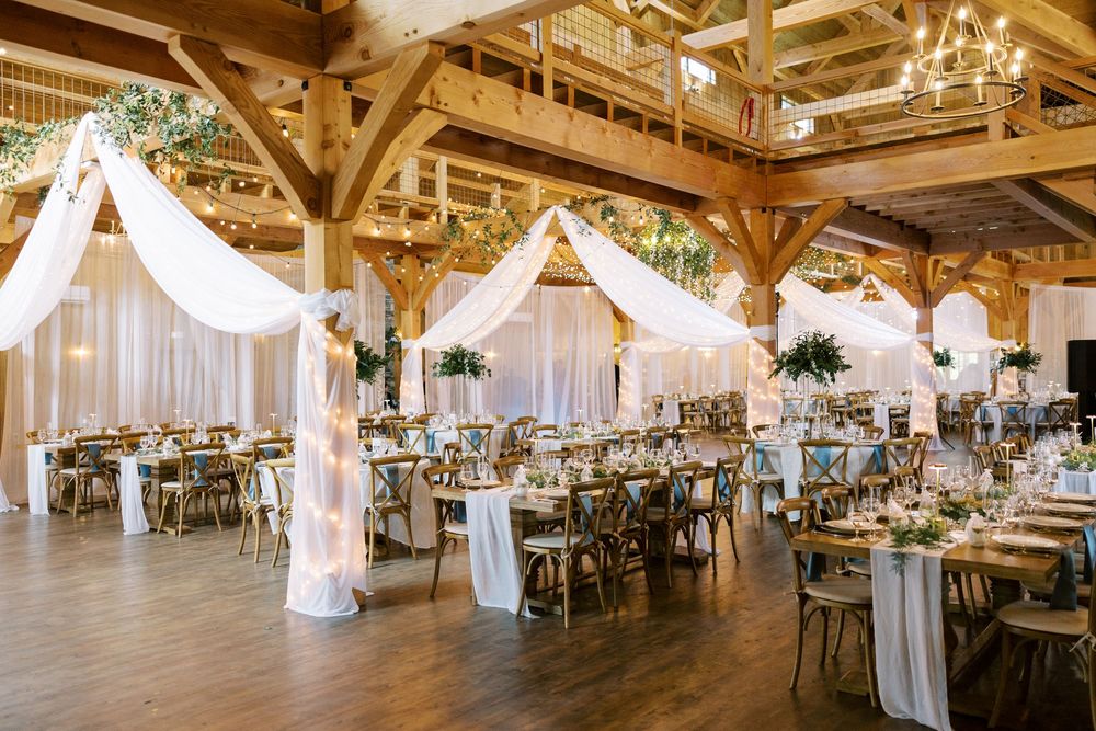 Elegant wedding reception venue with white draped canopies, rustic wooden beams, and gray table settings in a barn at Windham Manor.