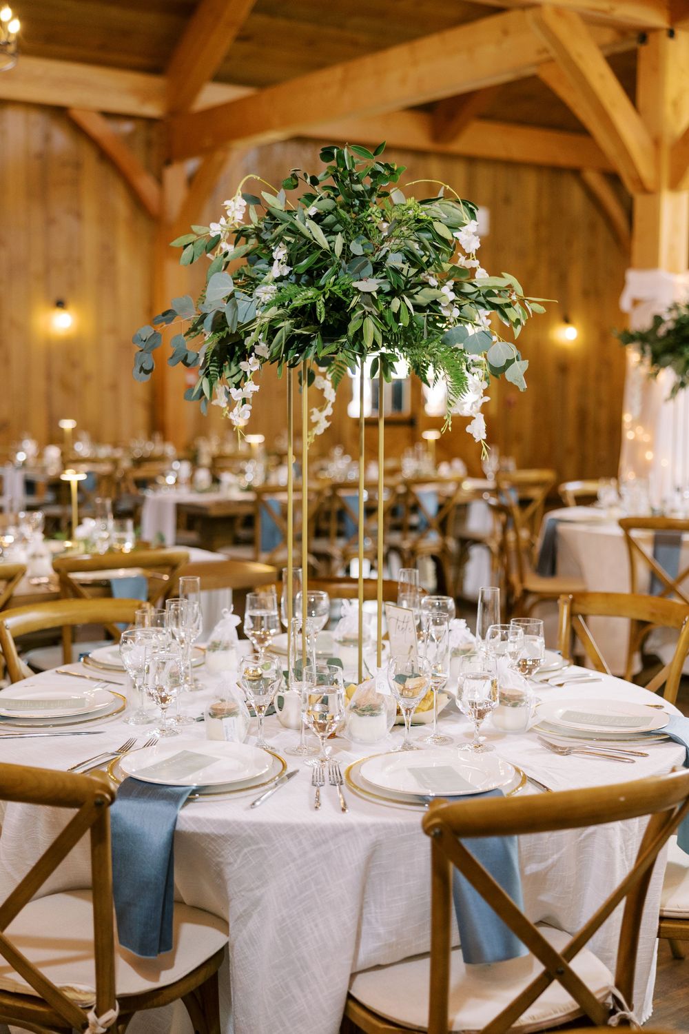 Elegant wedding reception table with tall greenery centerpiece, white linens, and rustic wooden chairs in a barn venue at Windham Manor.