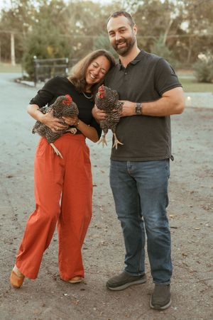 A joyful couple in casual attire holds brown chickens while standing outdoors at dusk.