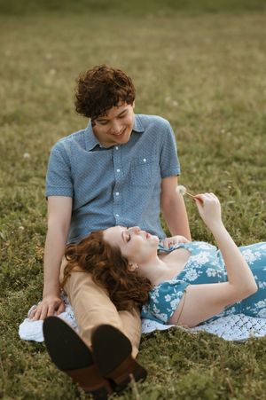 A couple relaxes on a blanket in a grassy field during a sunny day outdoors.