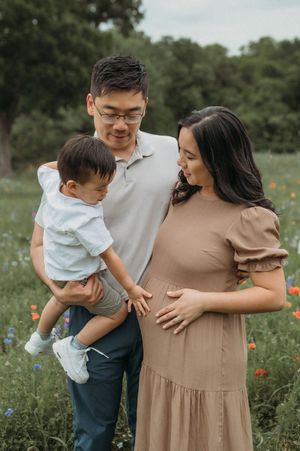 Expecting family in tan dress and white shirt posing together in a wildflower field during sunset.