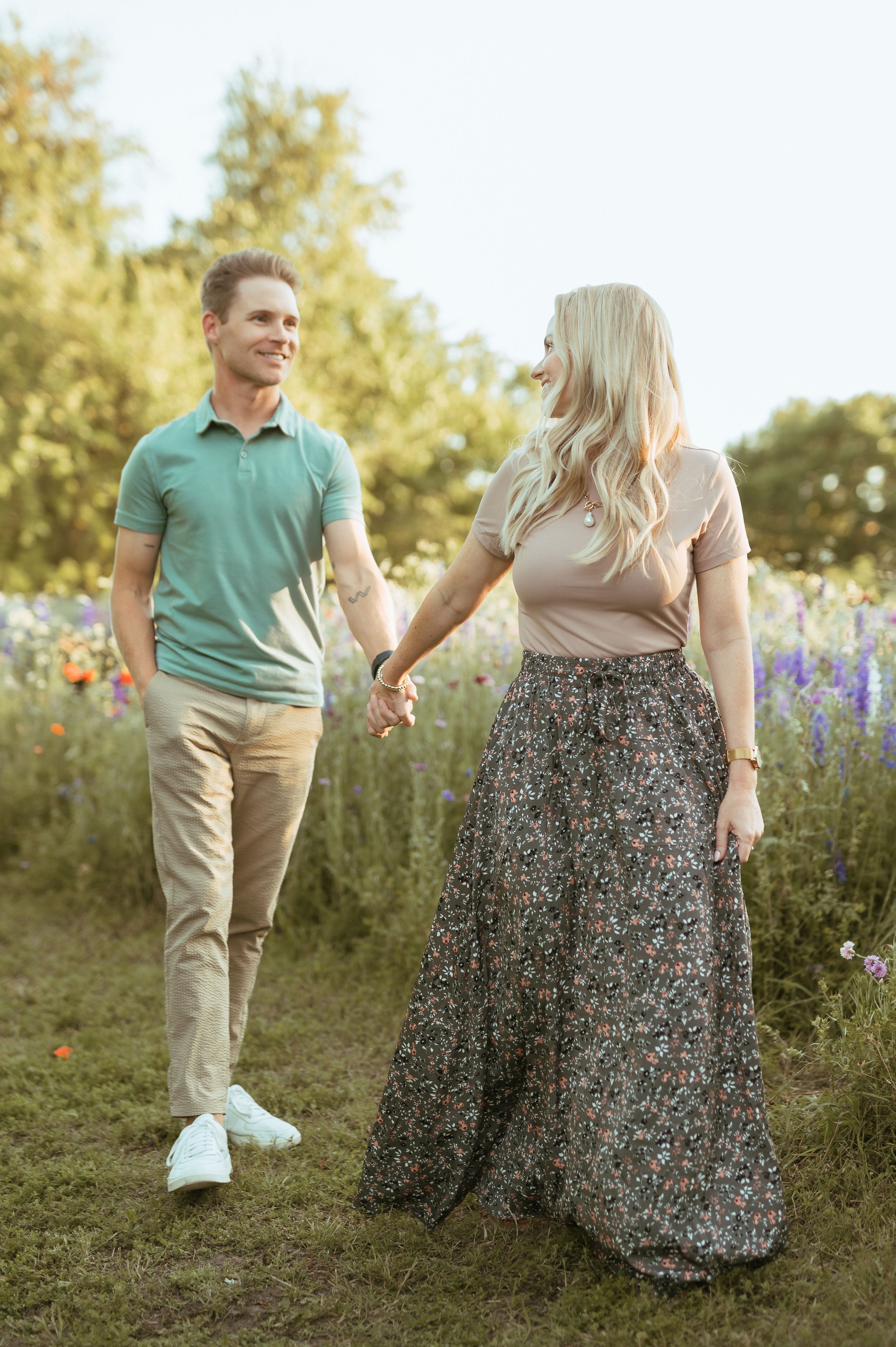 A couple holds hands while walking through a wildflower field at golden hour in casual summer attire.
