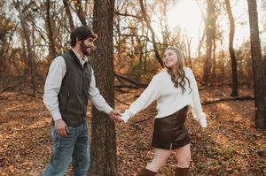 A joyful couple holds hands and smiles while walking through a sunlit autumn forest with fallen leaves on the ground.