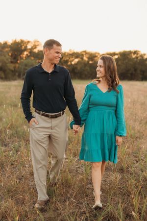 Couple walking hand in hand through a golden field at sunset, wearing casual attire of black and teal.