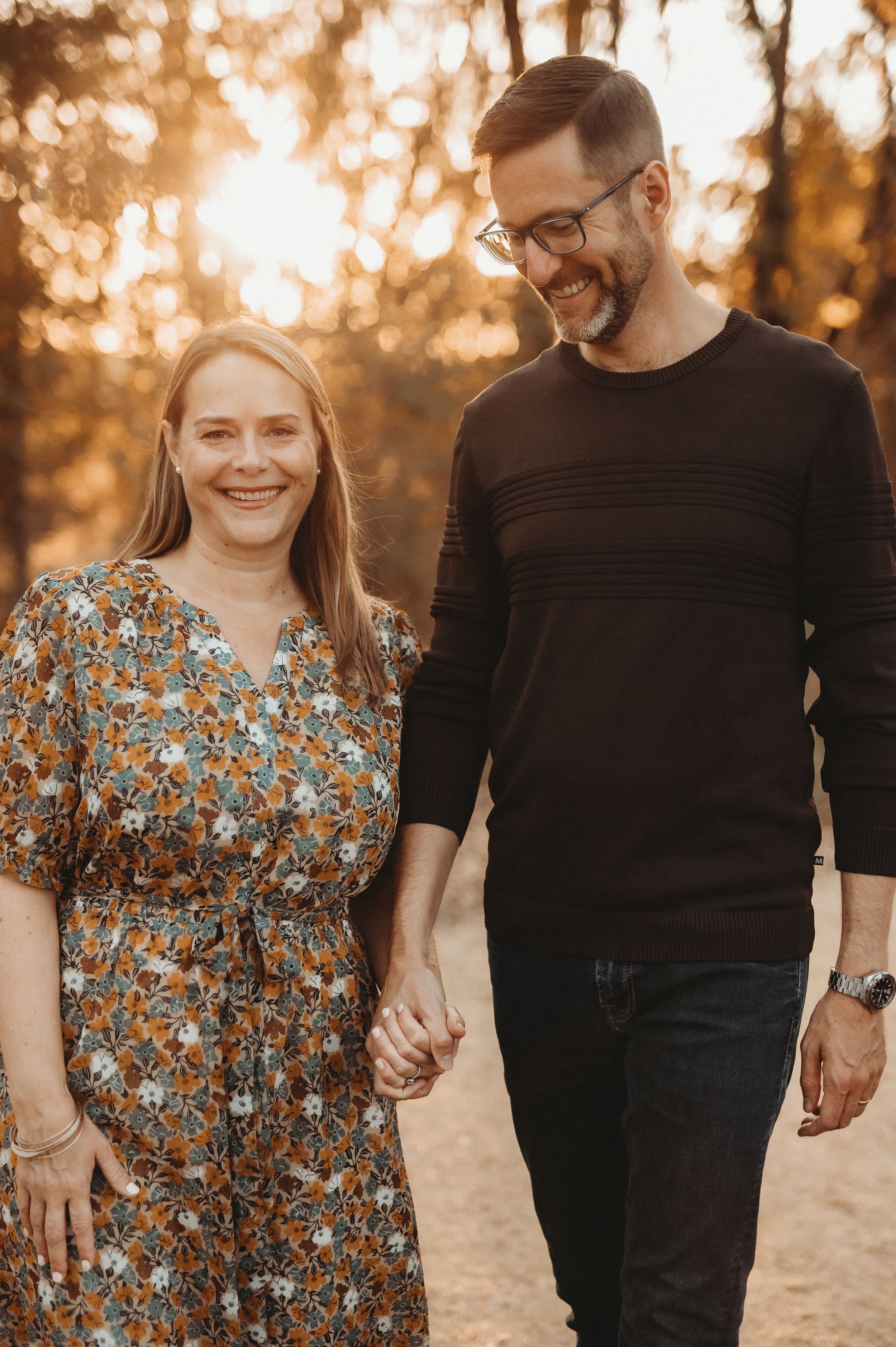 A couple walks hand in hand in autumn sunlight wearing casual attire against a warm golden background.