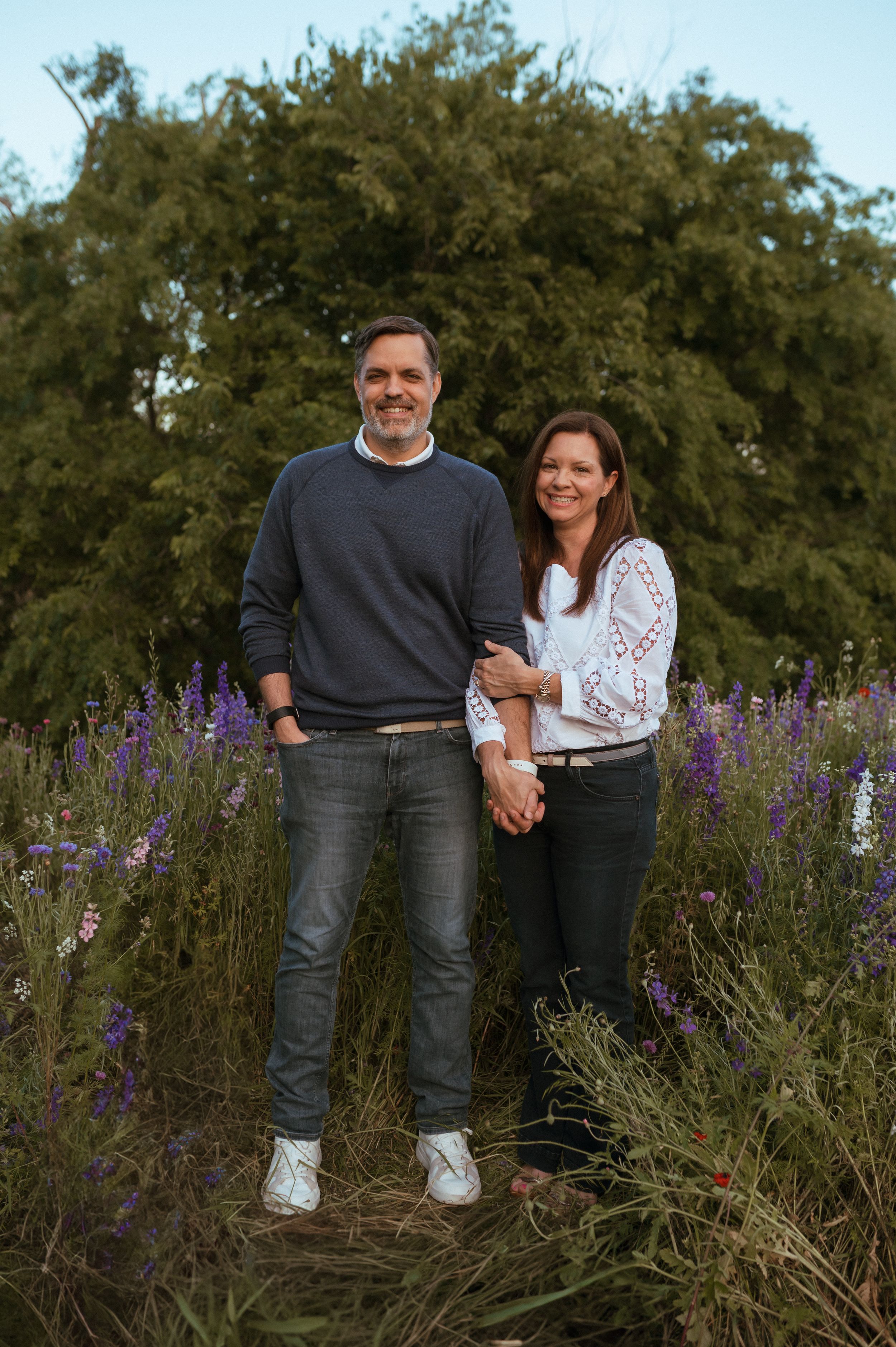 A couple stands together in casual attire among purple wildflowers and green foliage during a sunset photo session.