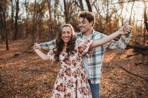 Two people in autumn clothing pose playfully on fallen leaves in a forest during golden hour sunset.