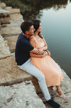 A couple sits together on rocky cliffs overlooking a serene lake during sunset in romantic poses.