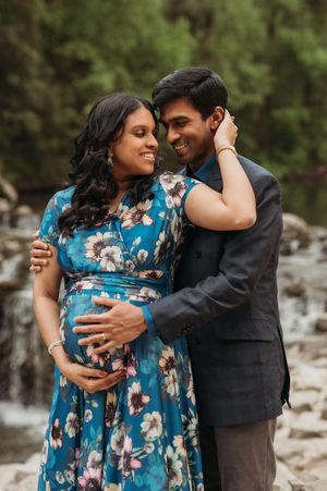 A couple shares an intimate moment by a waterfall, with a pregnant woman wearing a blue floral dress and person in dark suit.