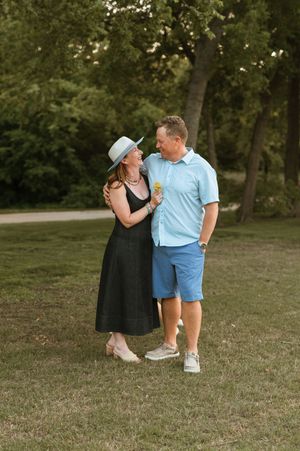 A couple in casual summer attire shares a romantic moment together on a grassy lawn surrounded by trees.
