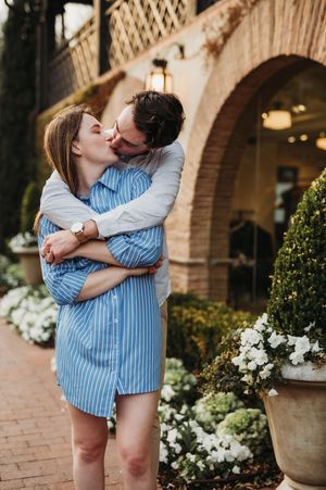 A couple embraces and shares a romantic moment in front of an elegant brick archway surrounded by flowering plants.