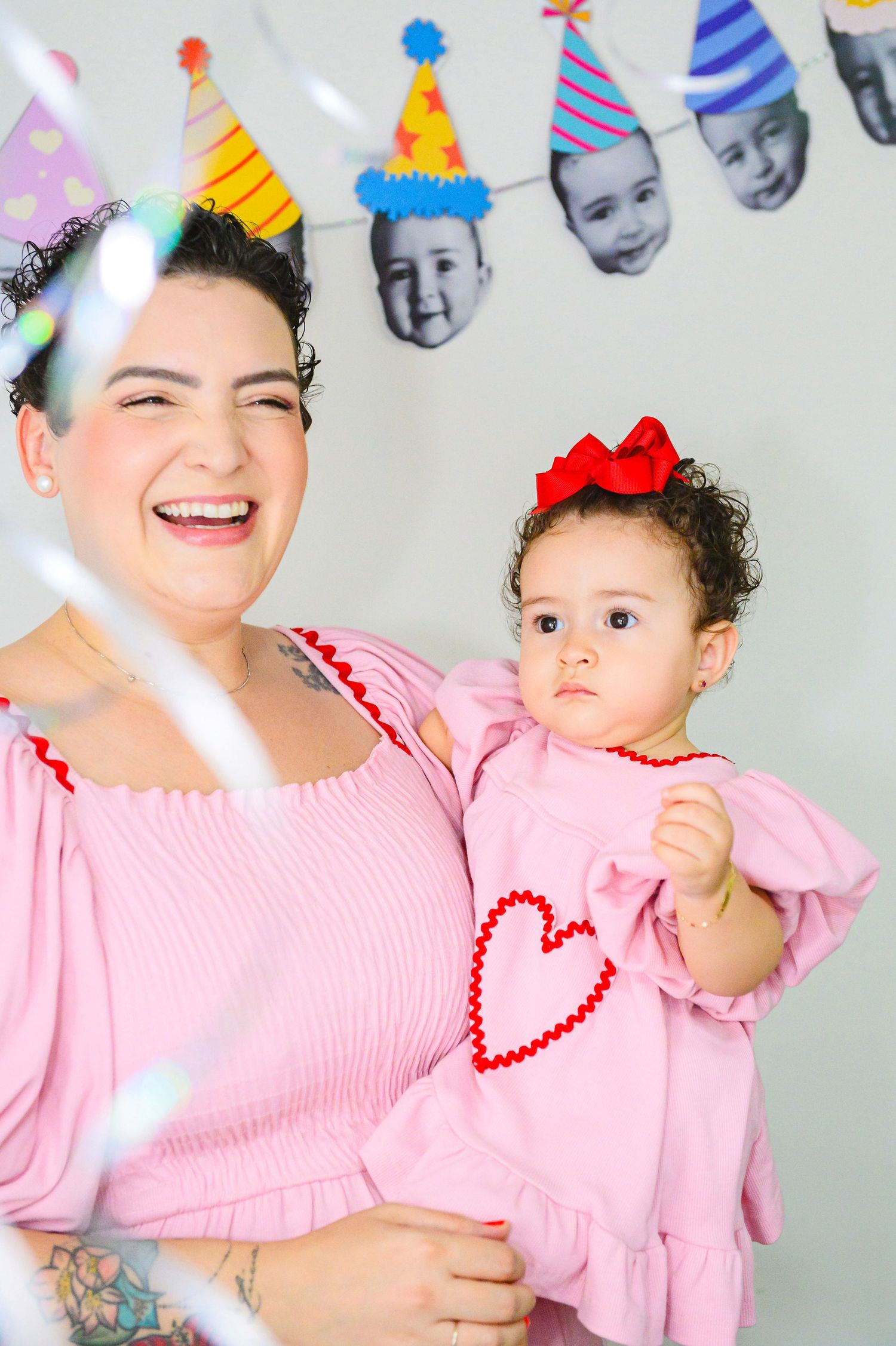Two people in matching pink outfits smile and celebrate at a party with decorative party hat banners in the background.