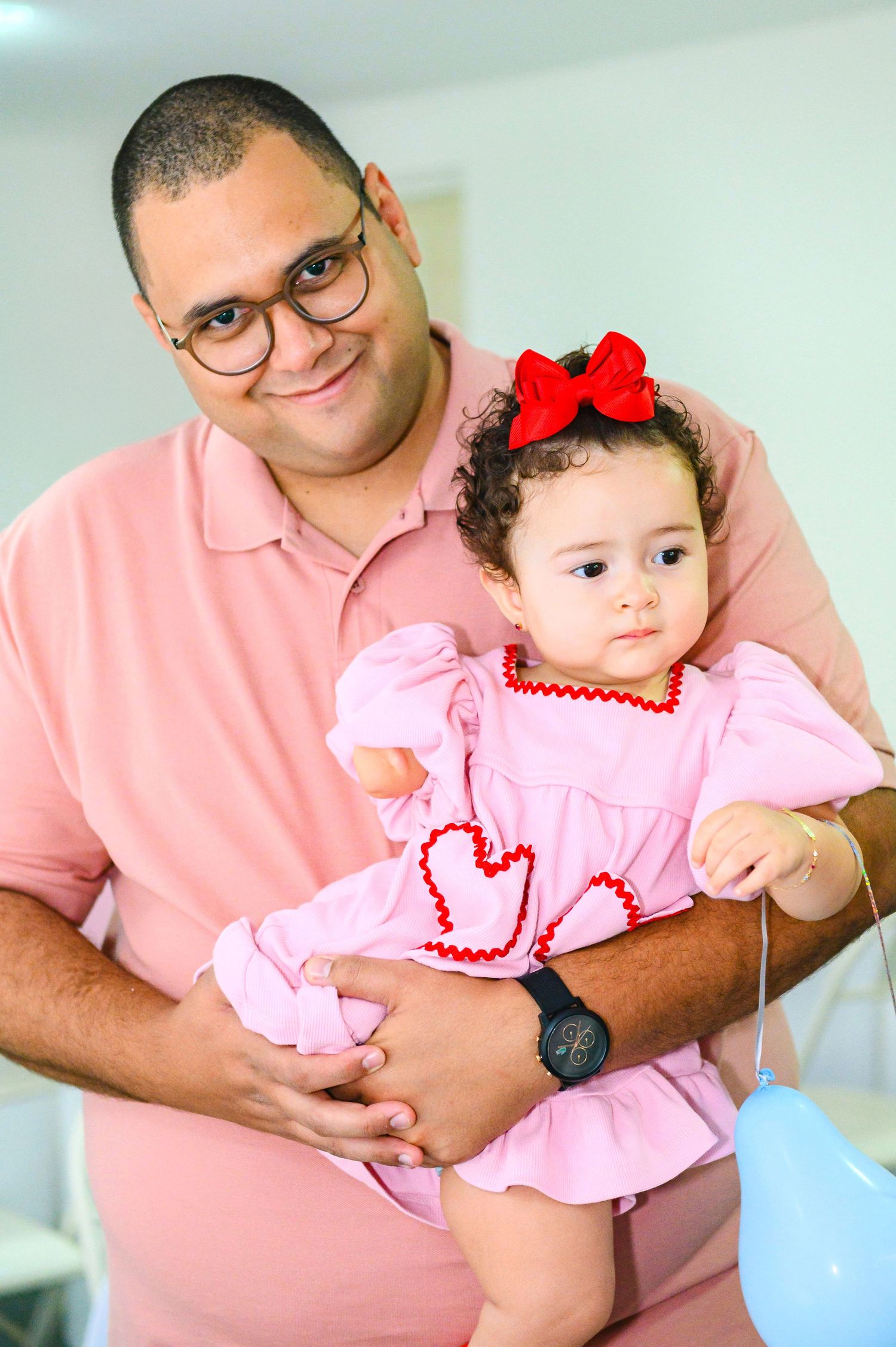 A gentle moment captured as a person in a pink polo shirt holds a baby wearing a pink dress with a red bow.
