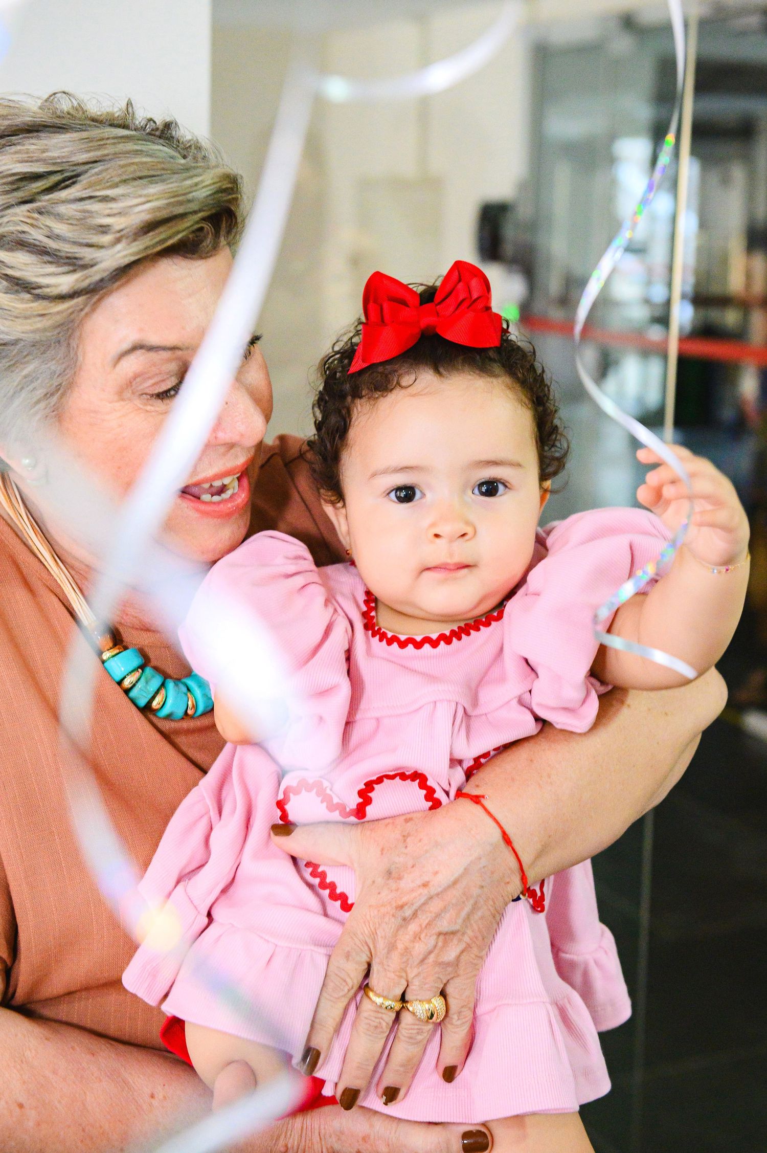 A small child in a pink dress with a red bow plays with streamers while being held at a celebration.