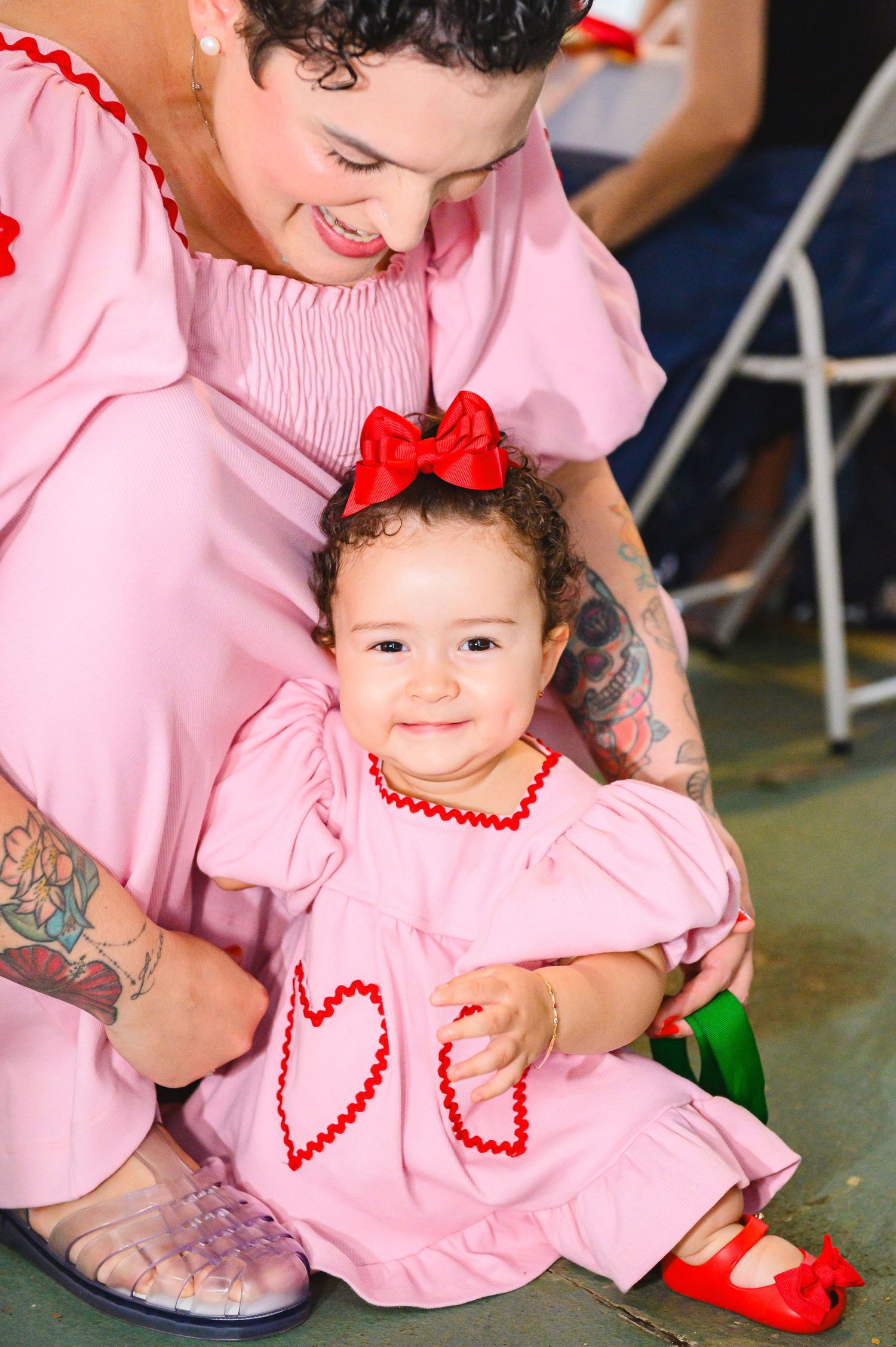 Parent and baby in matching pink outfits with red accents smile together at a special occasion.