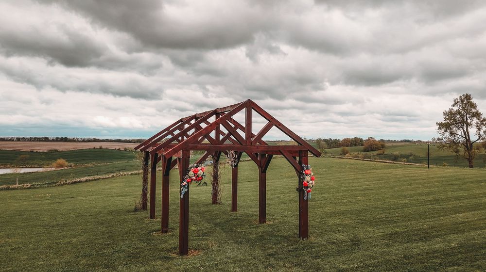 A rustic wooden pergola structure stands in a green field under dramatic cloudy skies with decorative floral accents.