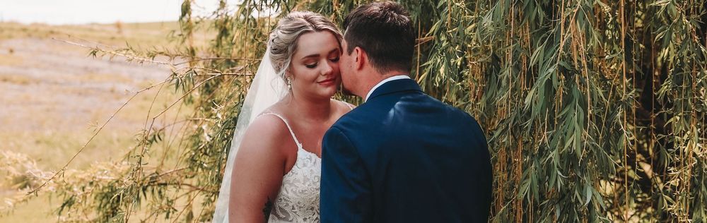 A wedding couple embraces among willow trees at golden hour in a scenic outdoor setting.