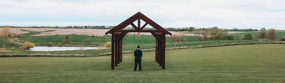 A person stands beneath a rustic wooden pergola structure overlooking a green landscape and pond.