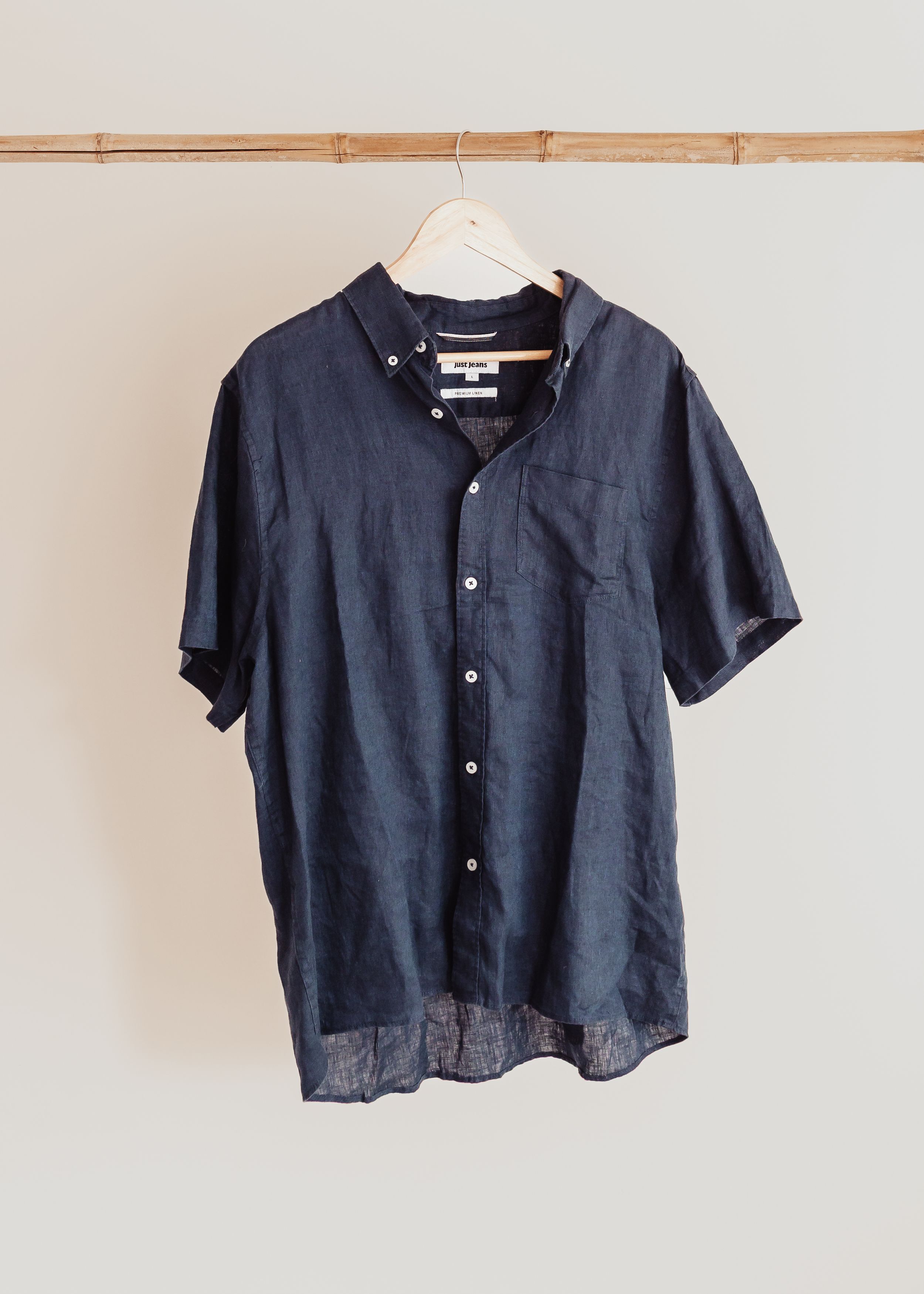 A navy blue short-sleeve button-down shirt with collar and chest pocket hanging on wooden rod against white wall.