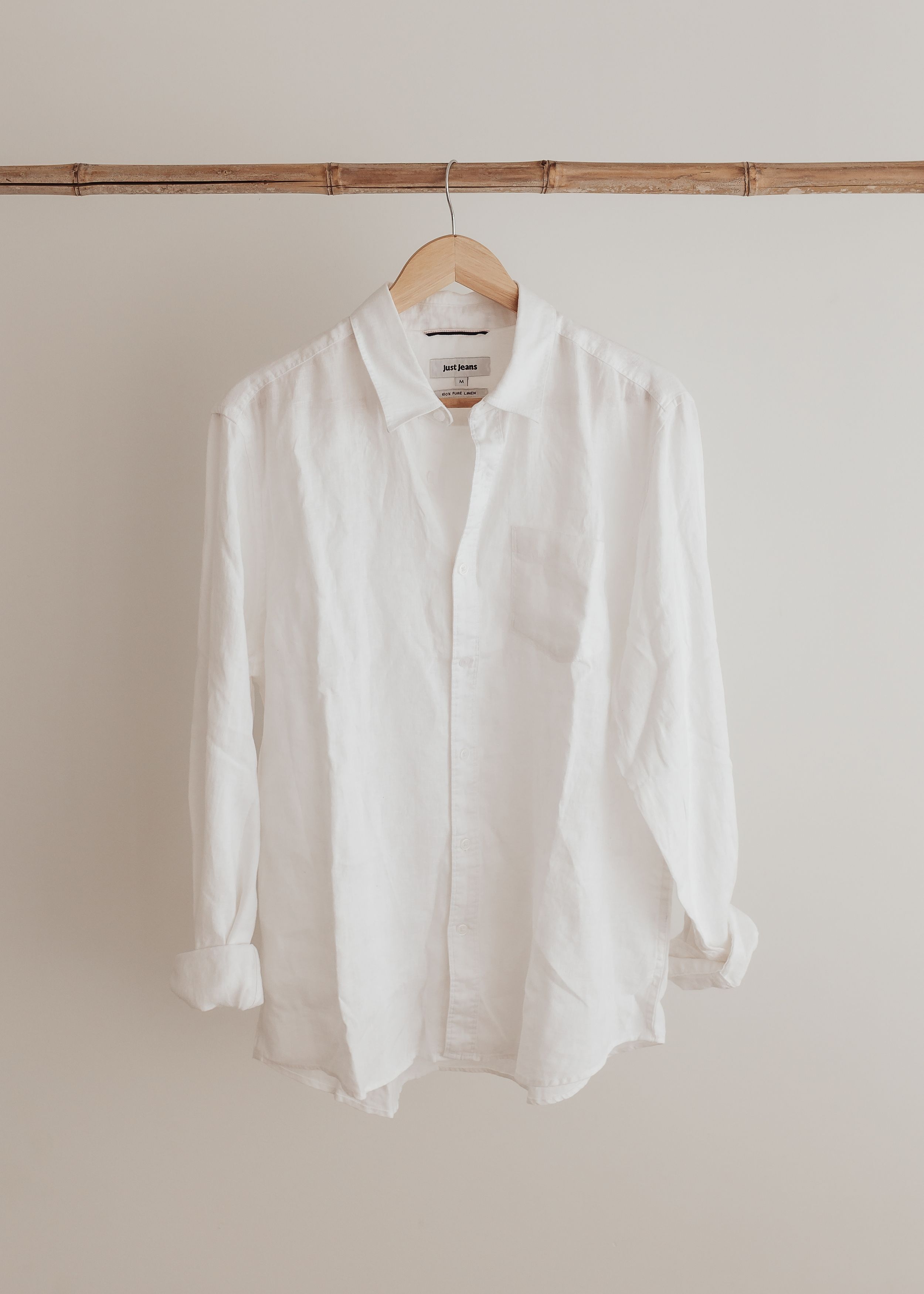 A crisp white linen button-down shirt hangs from a wooden hanger against a neutral beige wall.