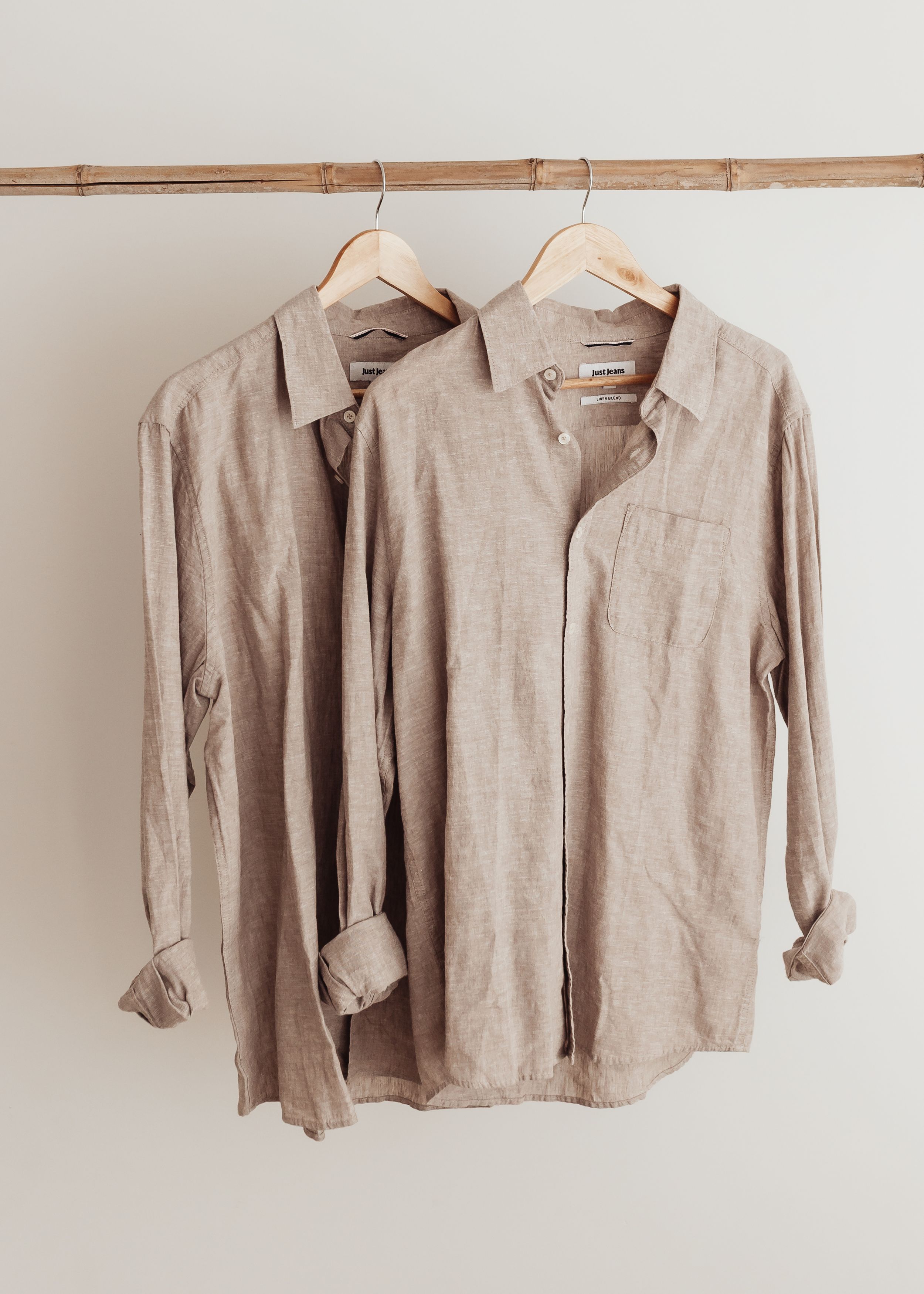 Two beige linen long-sleeve button-down shirts hanging on wooden rod against white wall.