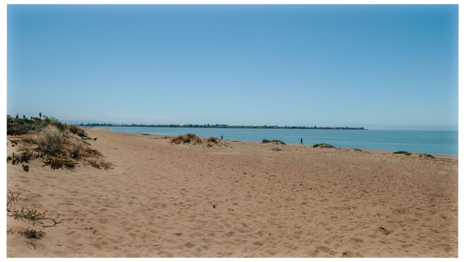 Sandy beach stretches along turquoise waters with dunes and distant shoreline under clear blue sky.