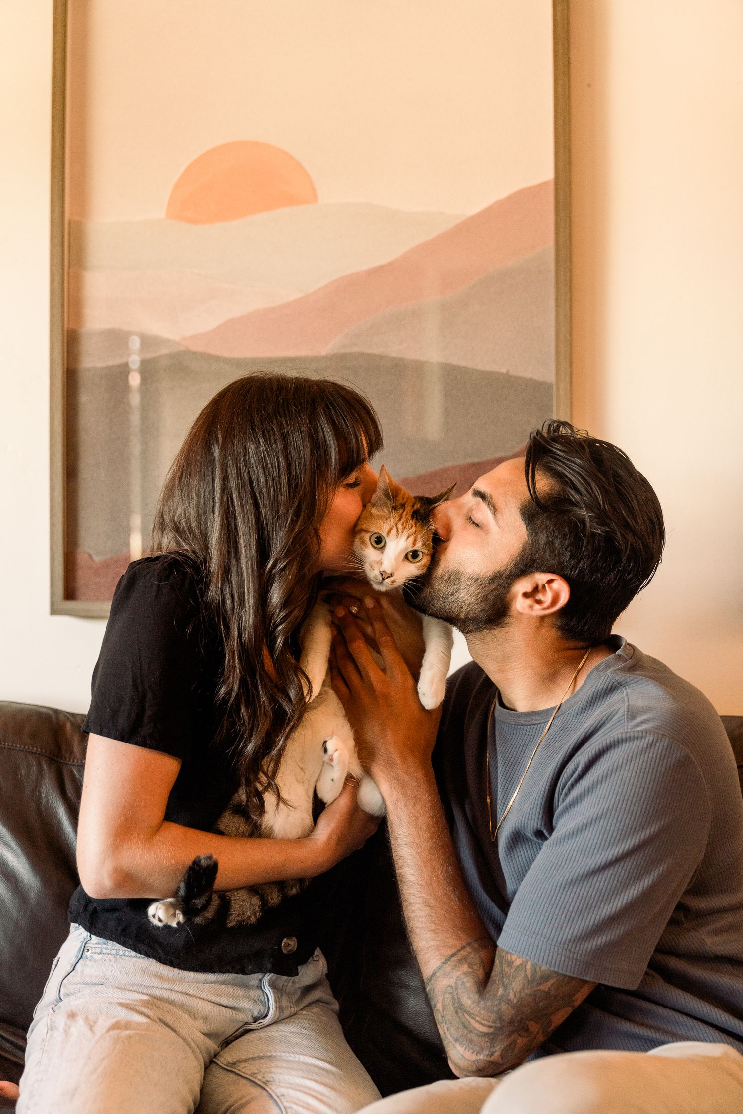 A loving couple kisses their ginger cat while sitting on a couch with a sunset painting in the background.