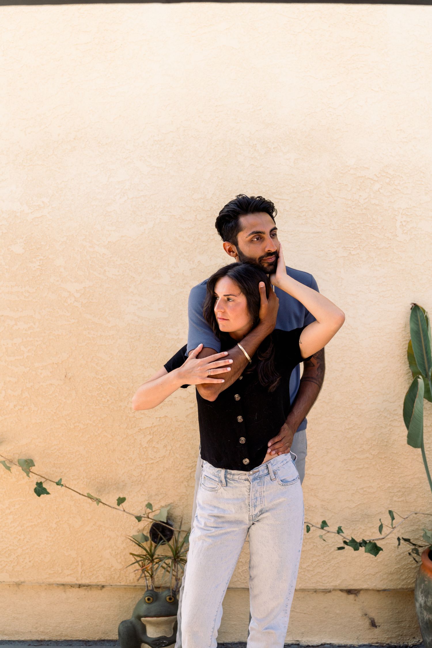 A couple embraces against a beige wall with a delicate vine plant growing in the background.