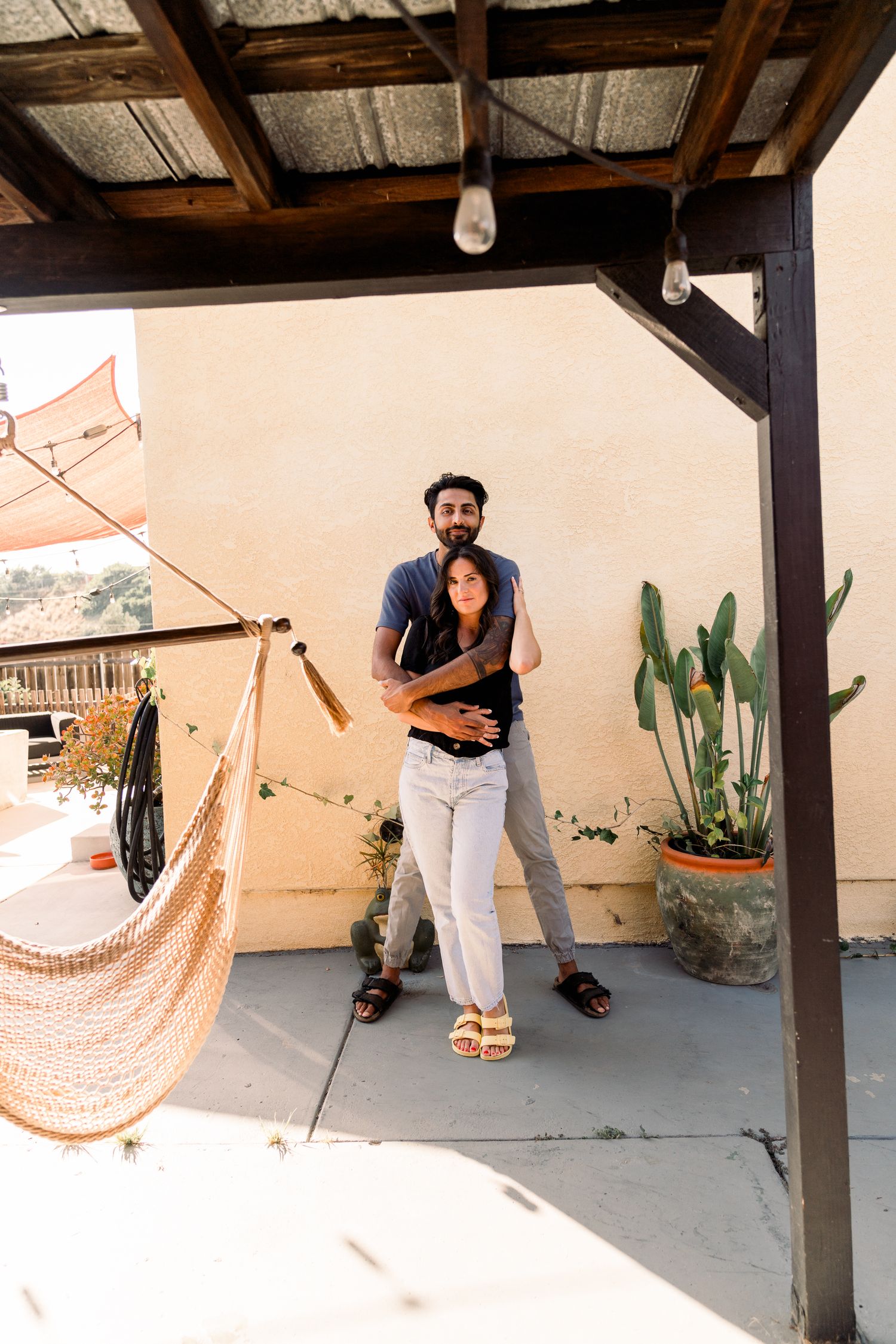A couple embraces on a patio with decorative string lights, hammock and potted plants in the background.