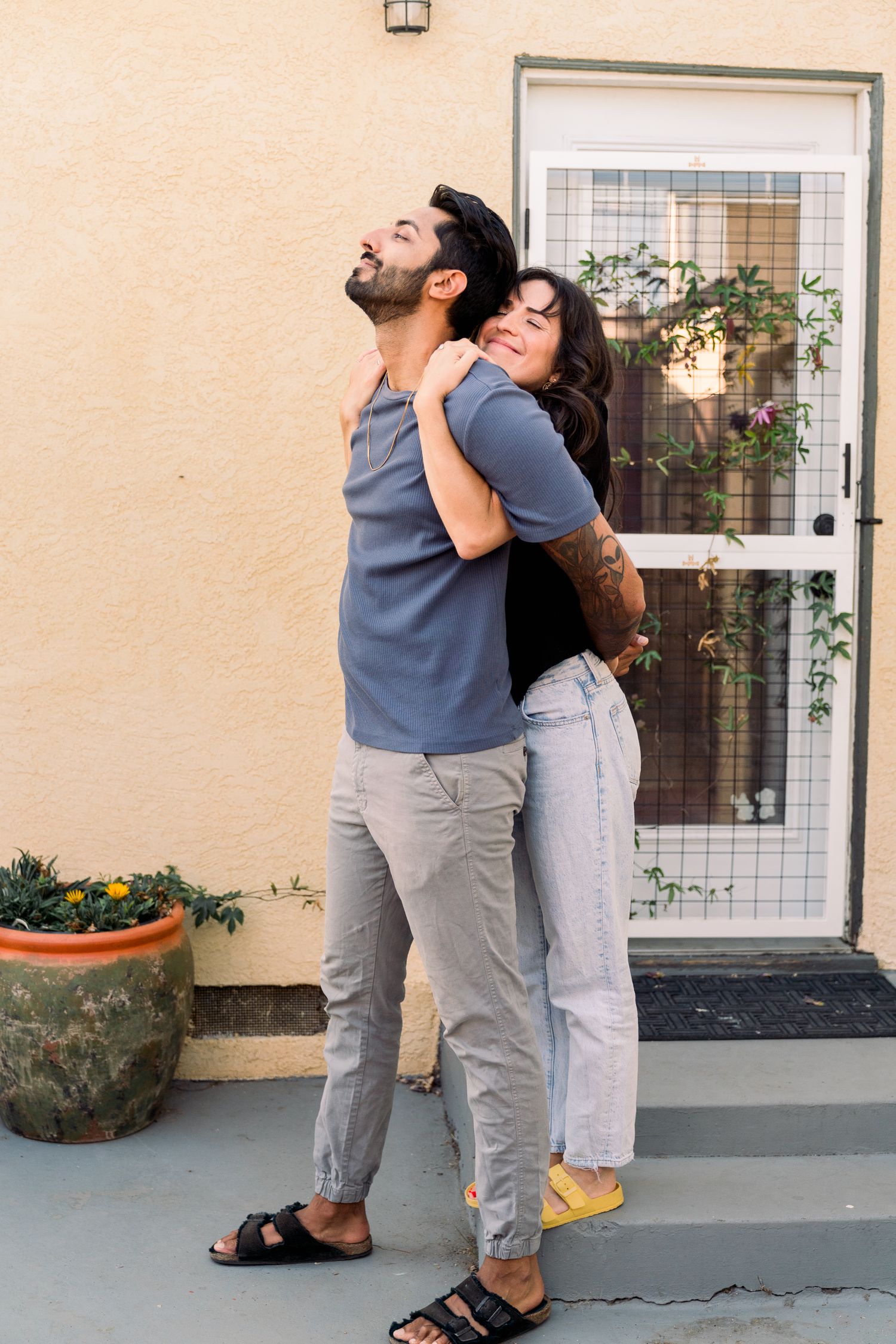 A couple shares an affectionate moment against a beige wall with a screened door and potted plants.