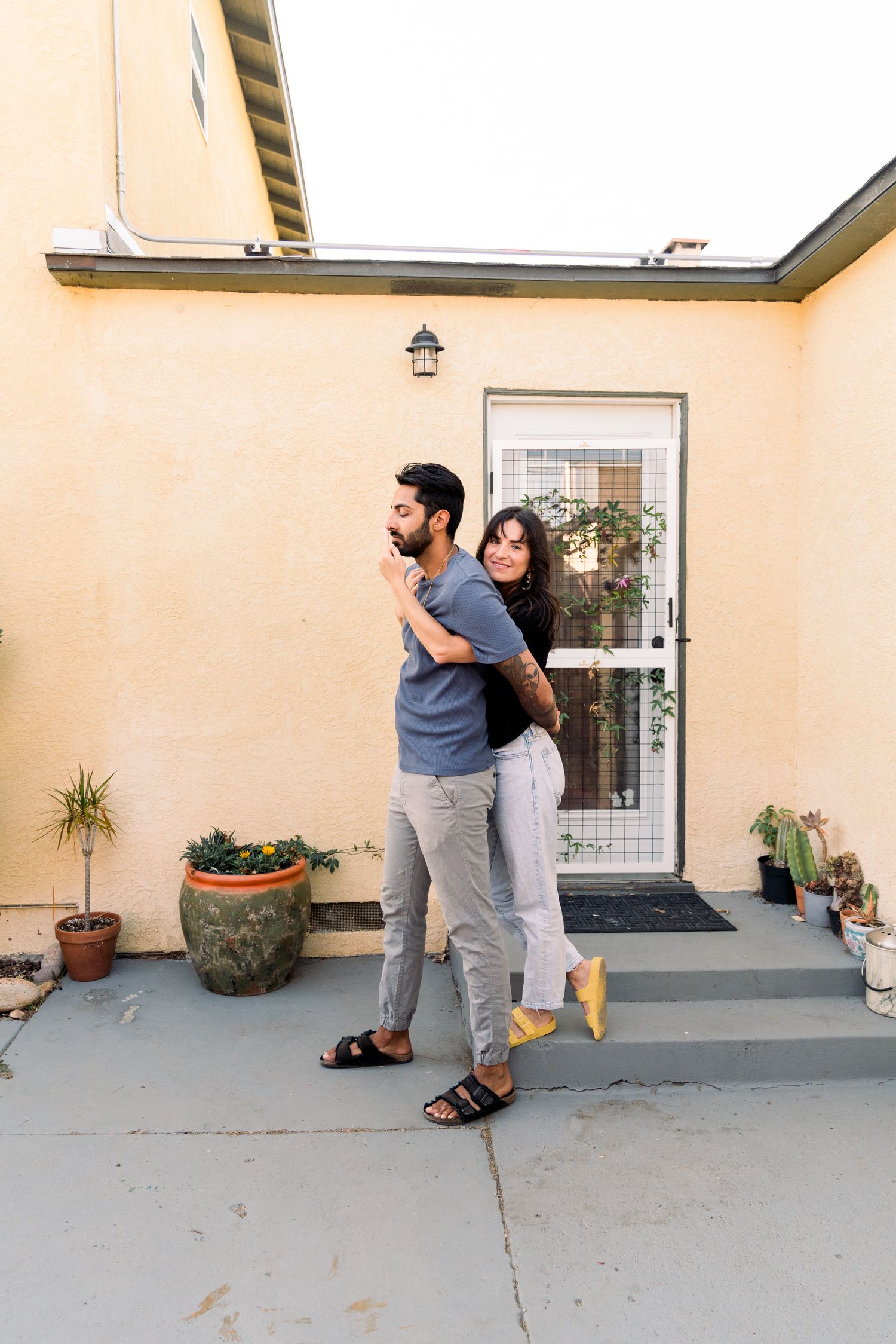 A couple embraces on an outdoor patio with potted plants and a screen door in the background.