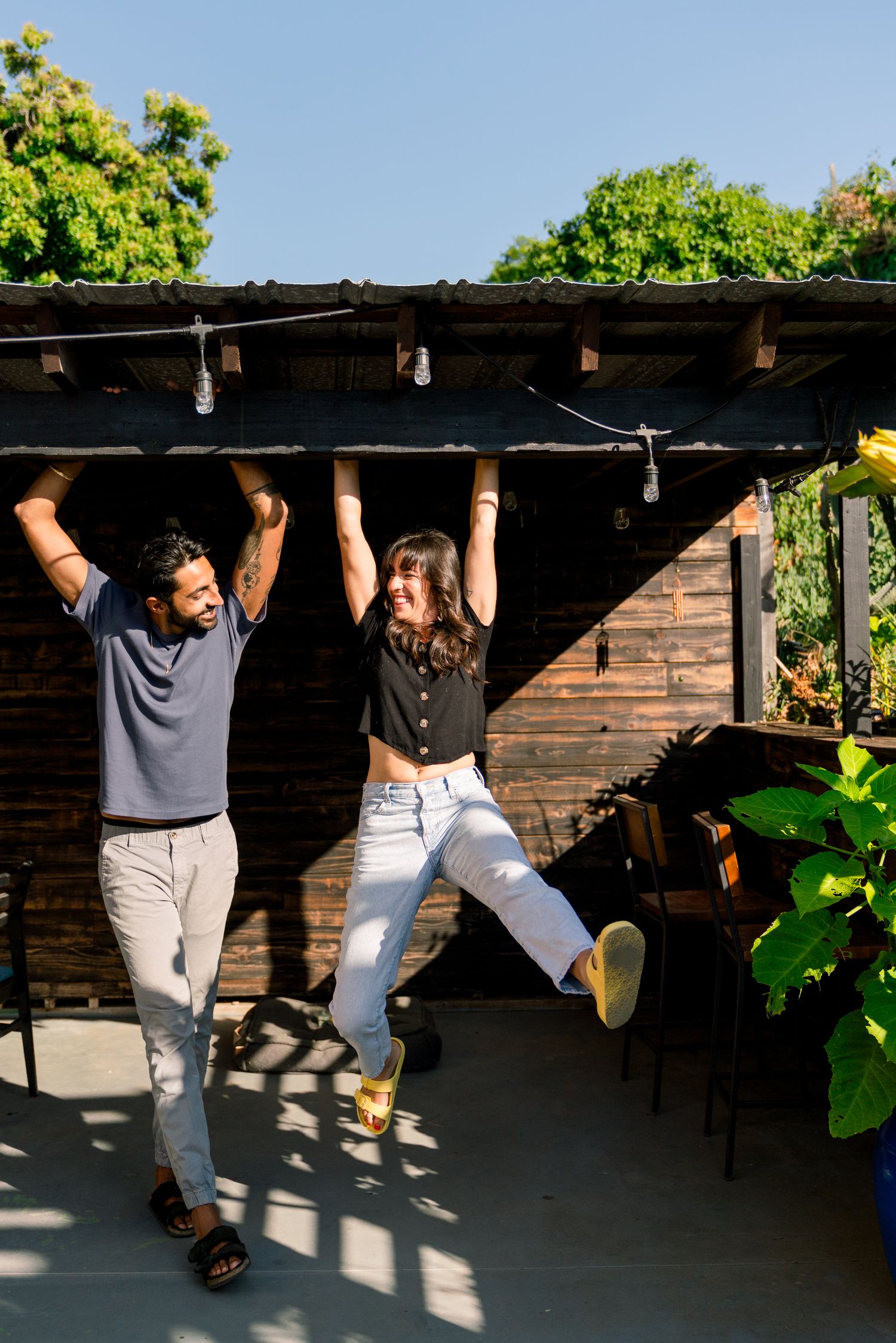 Two people playfully hanging from a wooden pergola structure in an outdoor patio setting.