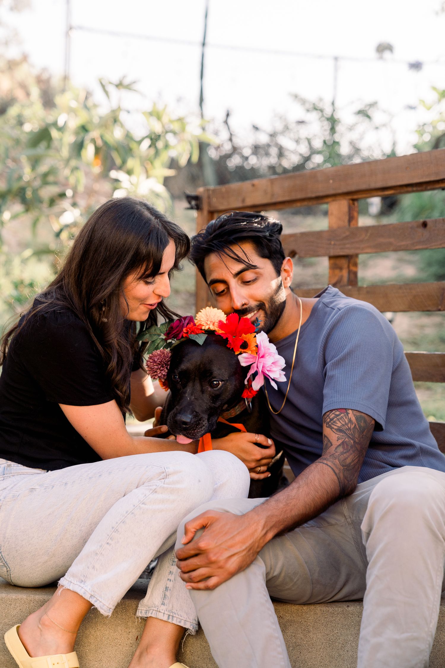 A couple sits together with their black dog wearing floral decorations in an outdoor setting.