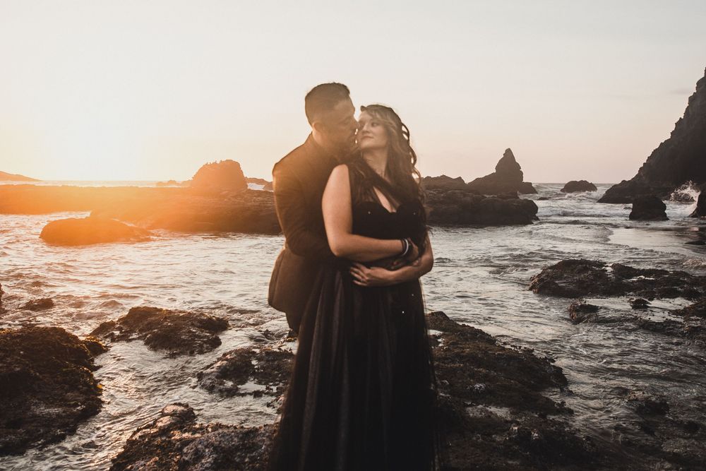 Silhouettes embrace on rocky coastline during golden hour with dramatic ocean and cliff backdrop.