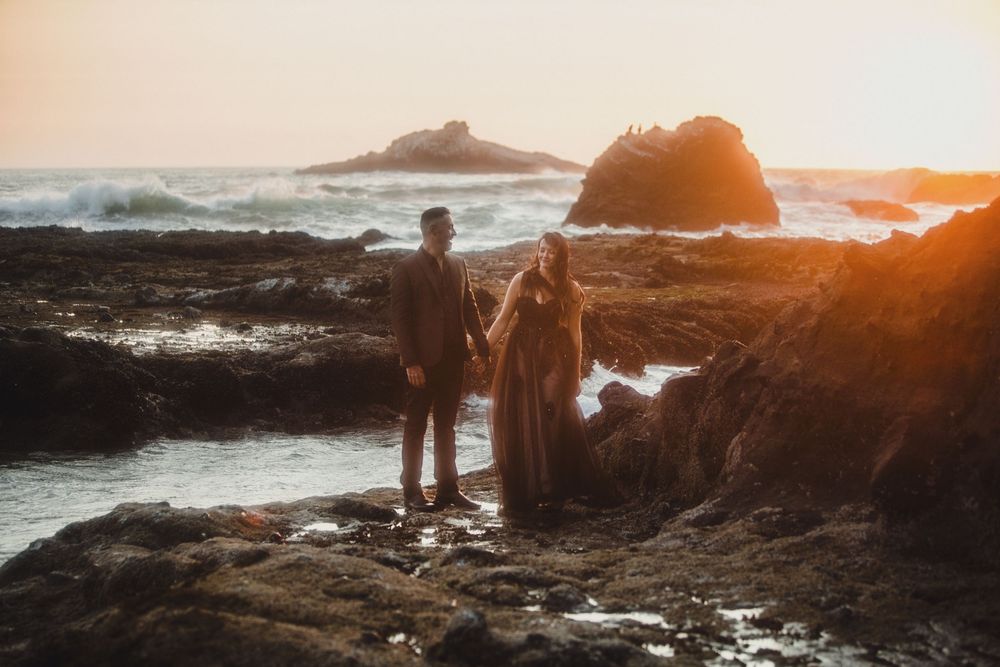 Couple holding hands while standing on rugged oceanside cliffs as sun sets over crashing waves.