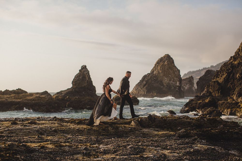 Two people walk together on rocky coastal cliffs with dramatic ocean waves and rugged sea stacks in Oregon.
