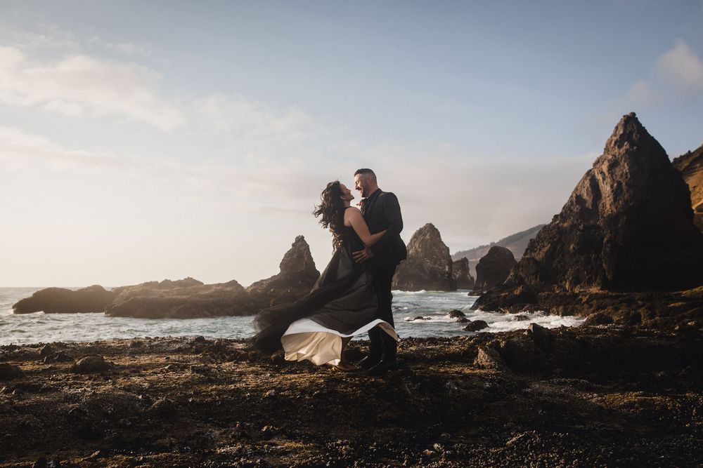 Two people embrace on rocky coastline with dramatic cliffs and crashing waves at sunset.