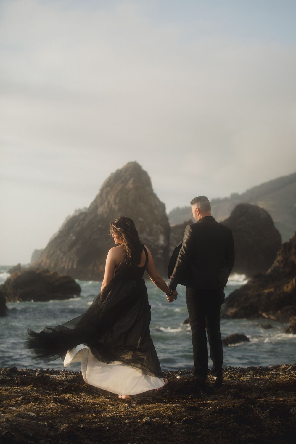 Couple in evening attire holding hands while standing on rocky coastal shore with dramatic ocean waves and cliffs.