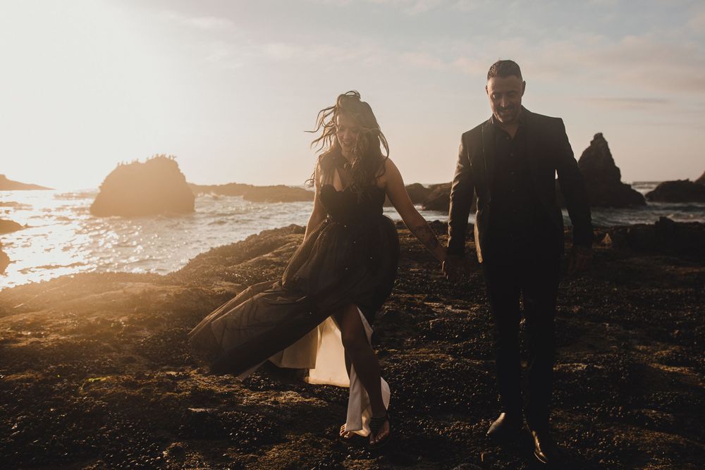 A silhouetted couple walks along a rocky beach at sunset with waves crashing against coastal rocks in the background.