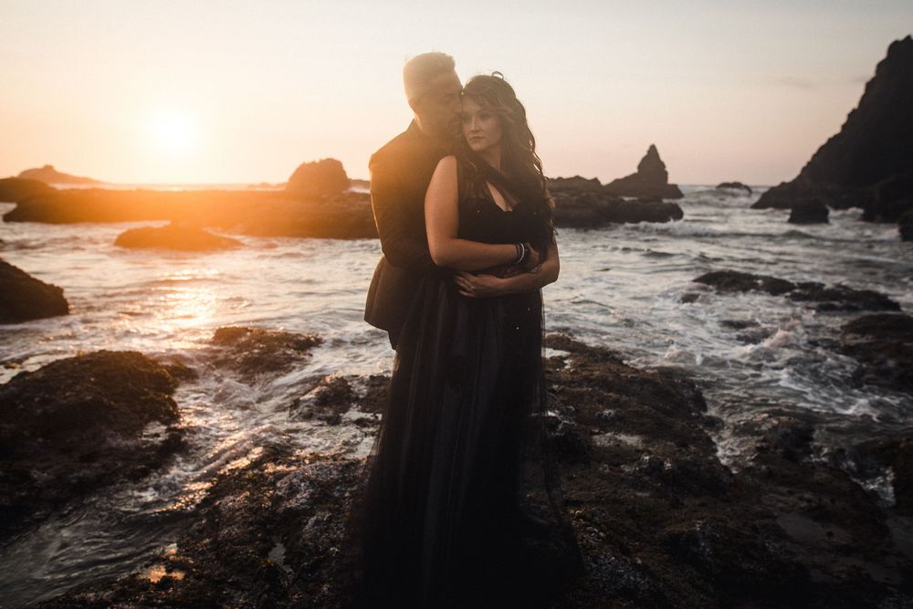 Silhouetted couple embraces on rocky beach at sunset with waves crashing against dramatic coastal rocks in background.