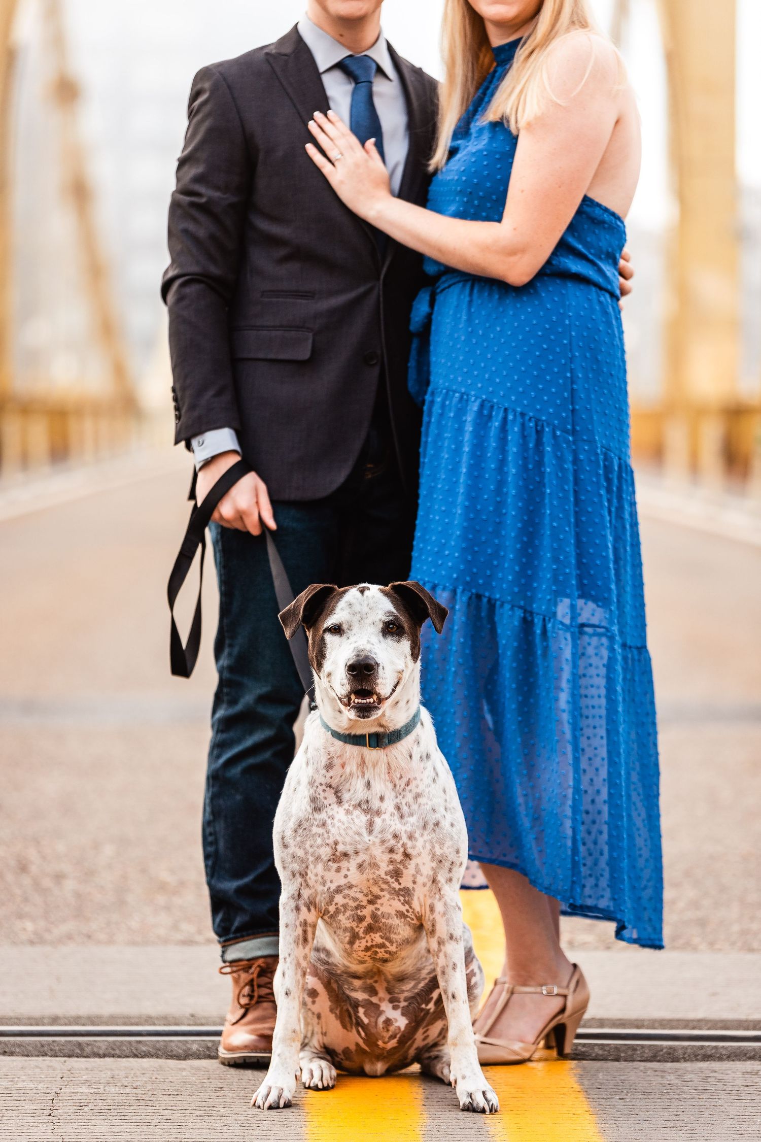 A spotted brown and white dog sits on a bridge while a couple in formal attire stands behind wearing a blue dress and suit.