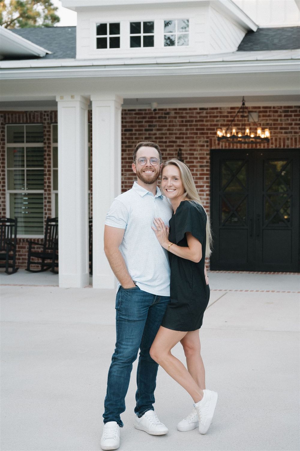 A couple stands together on a covered porch in front of a brick house with white trim and a dark front door.