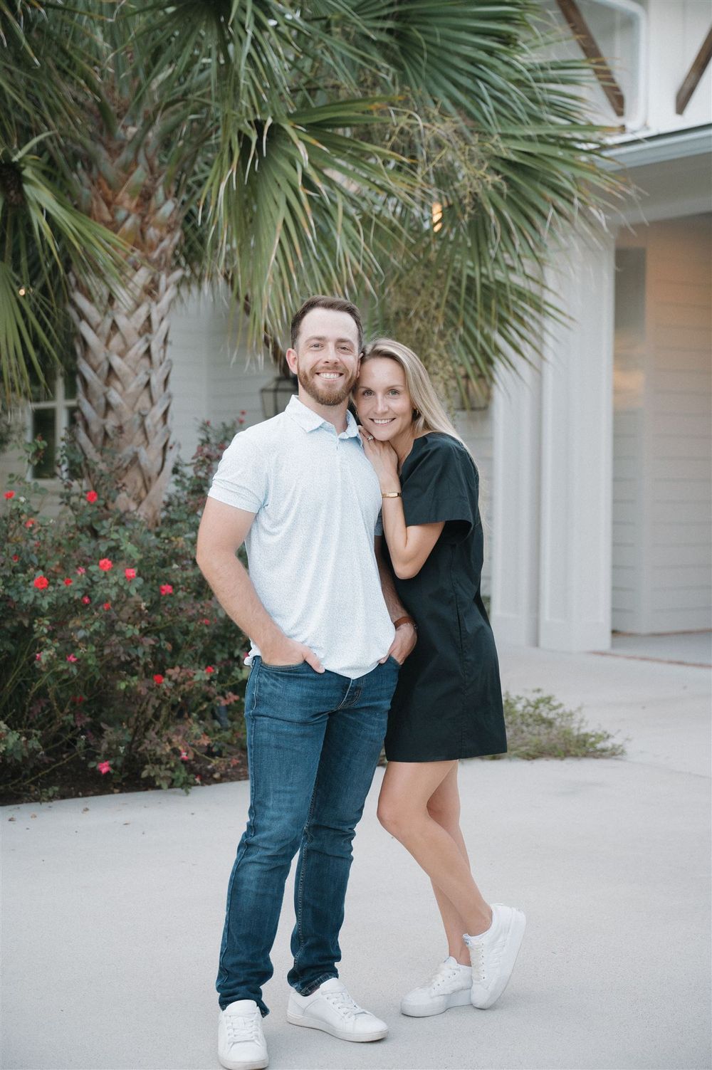 A couple poses together outdoors in casual attire next to a palm tree and white building exterior.