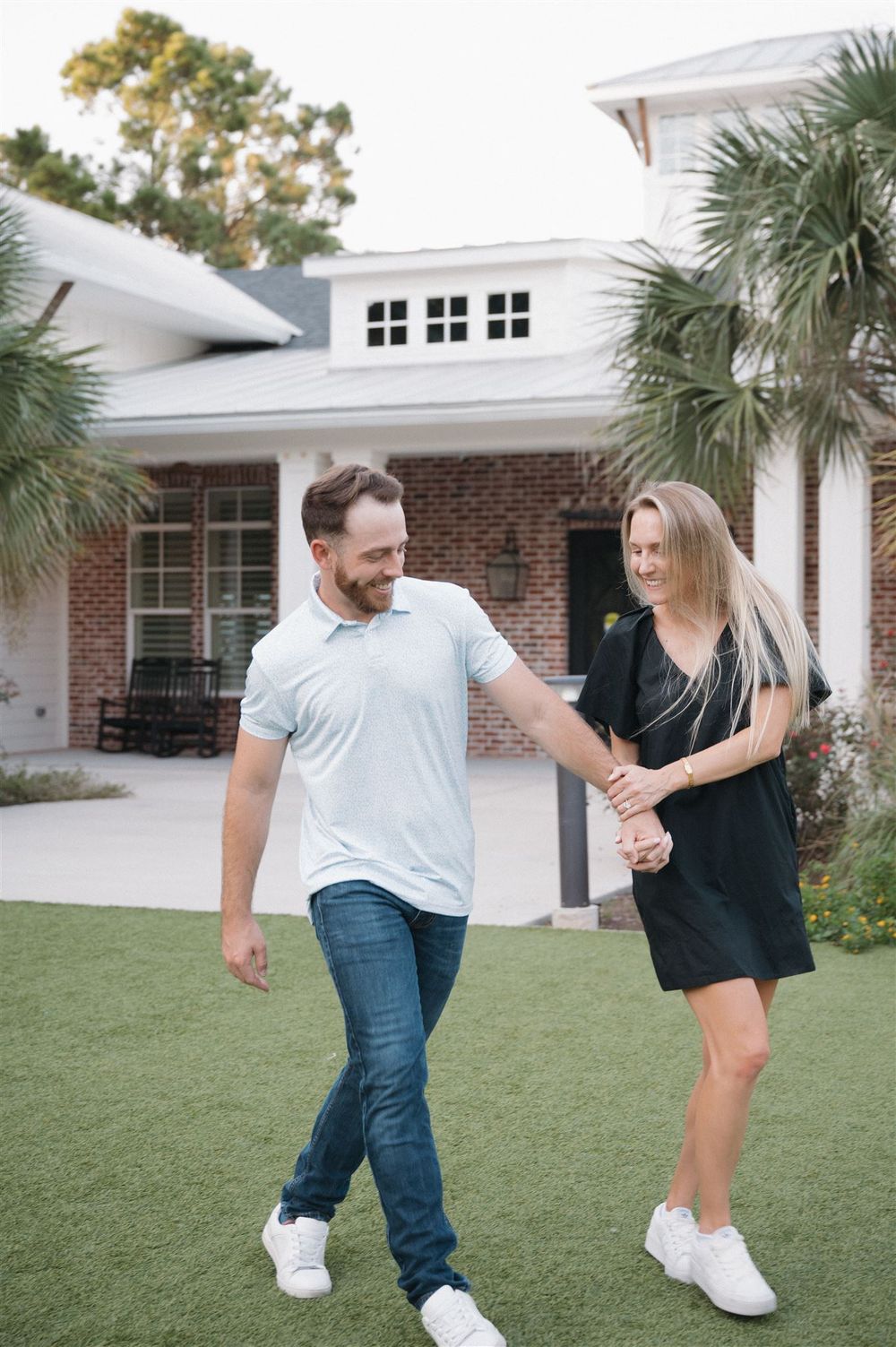 A playful couple shares a lighthearted moment together in front of a white brick house with palm trees.